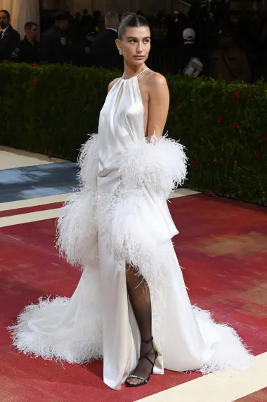 Hailey Bieber na ovogodišnju je Met Galu došla bez društva svog supruga Justina Biebera, a zasjala je u bijeloj haljini retro stila ukrašenoj bogatim nojevim perjem. Njezinu je haljinu dizajnirao Saint Laurent by Anthony Vaccarello, a napravljena je od najfinijeg bijelog satena. S dugačkim plaštem, odnosno ogrtačem, zračila je glamurom starog Hollywooda i prizivala u misli lookove 1920-ih godina. 